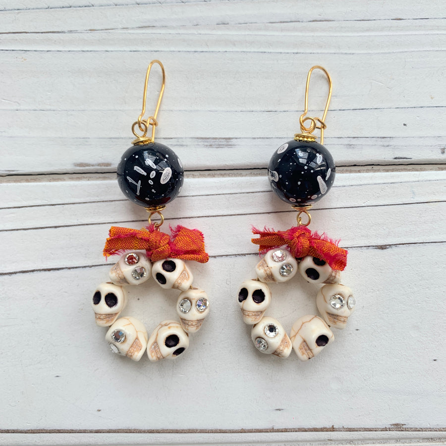 Lenora Dame Halloween earrings with white miniature carved sugar skulls, some with rhinestone embellished eyes, shaped into hoops and adorned with a bow made from orange and red sari silk. The skull hoops dangle from black with white splatter acrylic beads attached to gold-plated French hook ear wires.